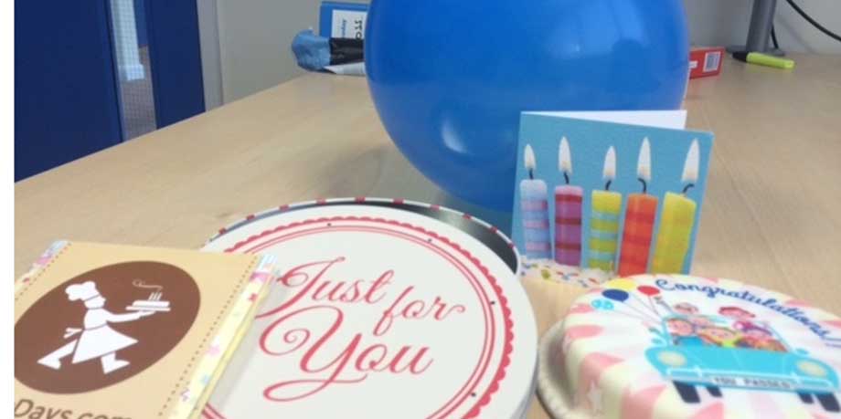 a photo of a small cake, blue balloon,  a birthday card, a cake tin and a leaflet from baker days